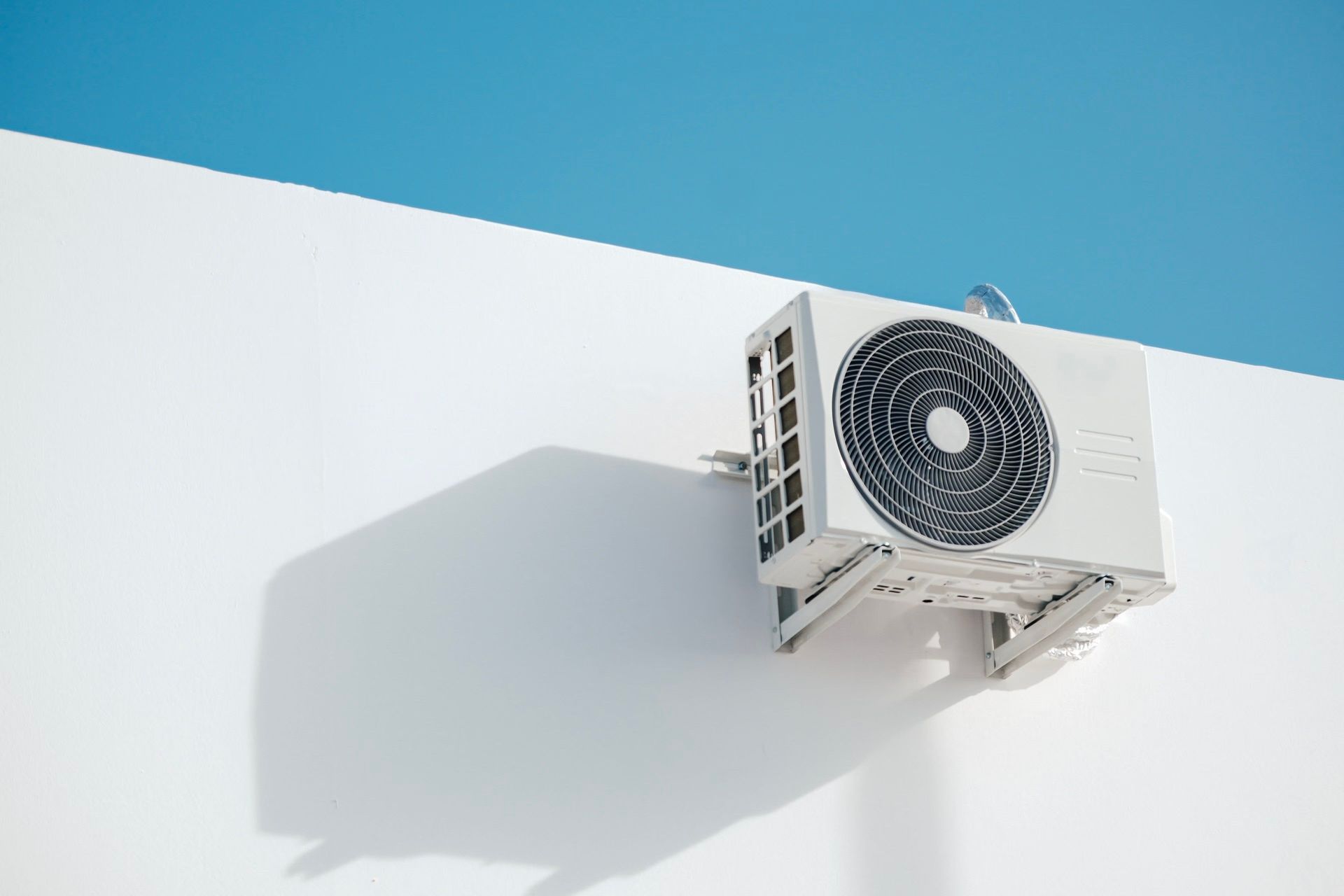 Wall-mounted air conditioner unit on a white building, set against a clear blue sky, creating a minimalist aesthetic.